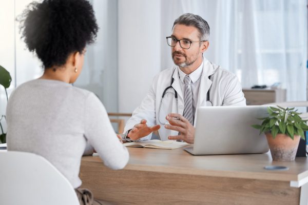 Een mannelijke arts voert een gesprek met een vrouw aan tafel. Hij heeft een doktersjas aan en stethoscoop om zijn nek. Op tafel staan een plant en laptop, en voor hem ligt een opengeklapt boek.